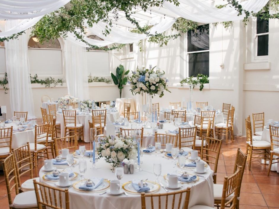 Elegant wedding reception setup with round tables, gold chairs, white linens, blue/white flowers, and hanging greenery under a canopy.