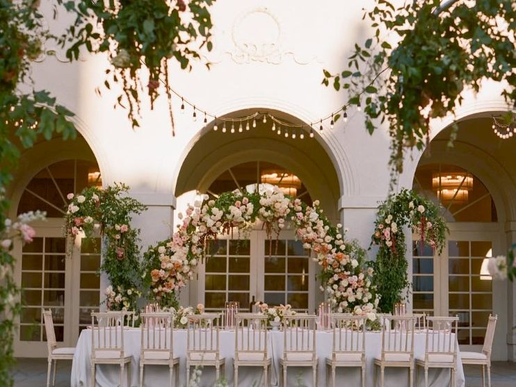 An outdoor wedding ceremony setup with white chairs, floral arches, and pastel bouquets under a decorated courtyard entrance.
