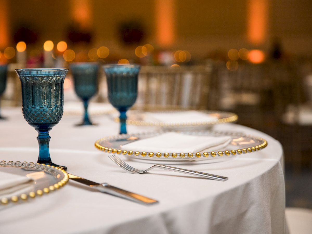 A banquet table set with teal blue goblets, gold beaded napkin rings, forks and plates, lit by warm bokeh lights.