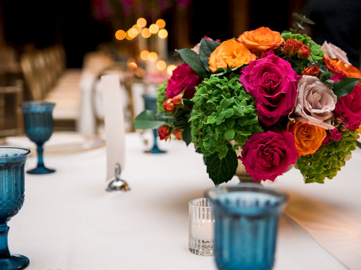 A lush bouquet of colorful roses and greenery sits on a reception table, with candles and bokeh lights glowing in the background.