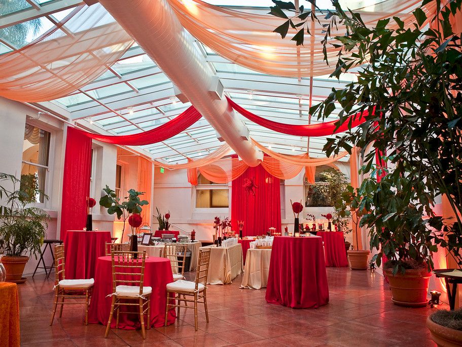 A festive indoor event setup with red-and-orange draped fabric, decorated banquet tables and chairs, and potted plants.