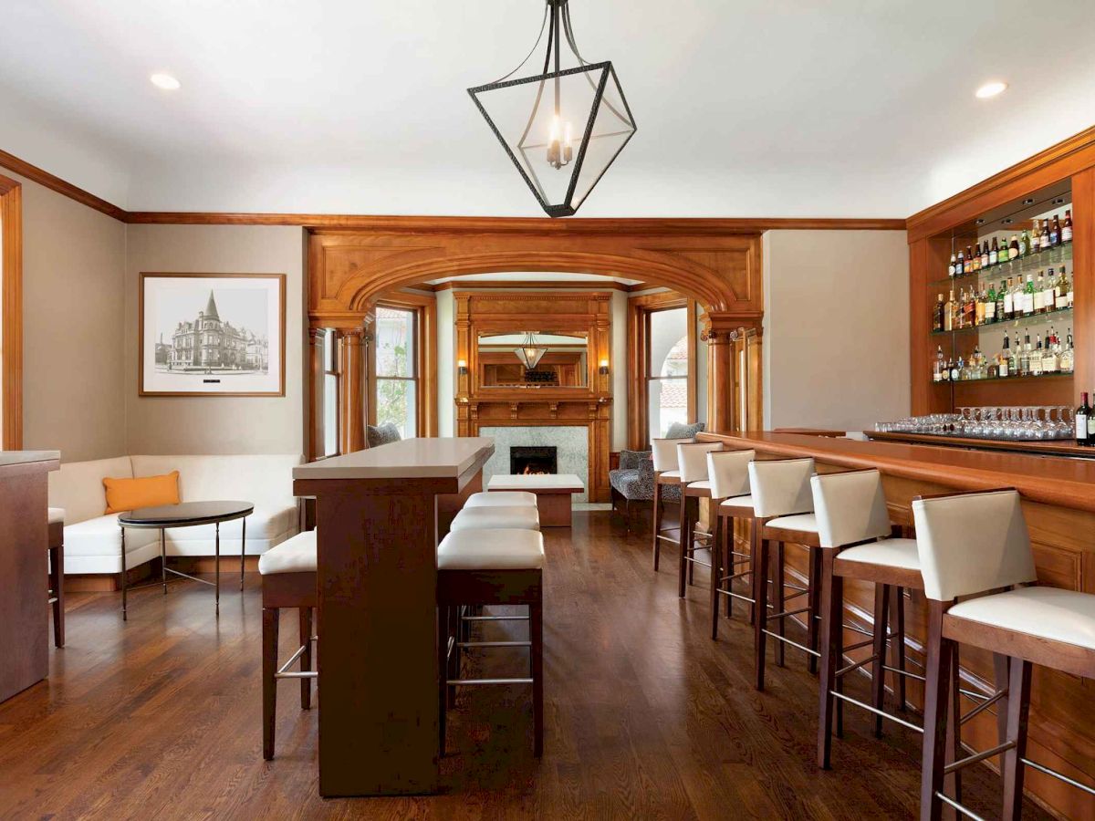 A stylish hotel bar with a long wooden counter, high stools, soft lighting, and a mirrored back wall; warm hardwood floors and seating area to the side.