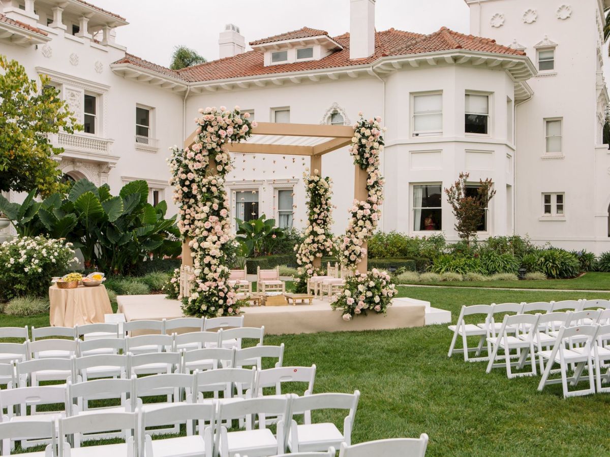 An outdoor wedding ceremony setup with white chairs, a floral arch, and a formal white villa backdrop, all decorated for the event.
