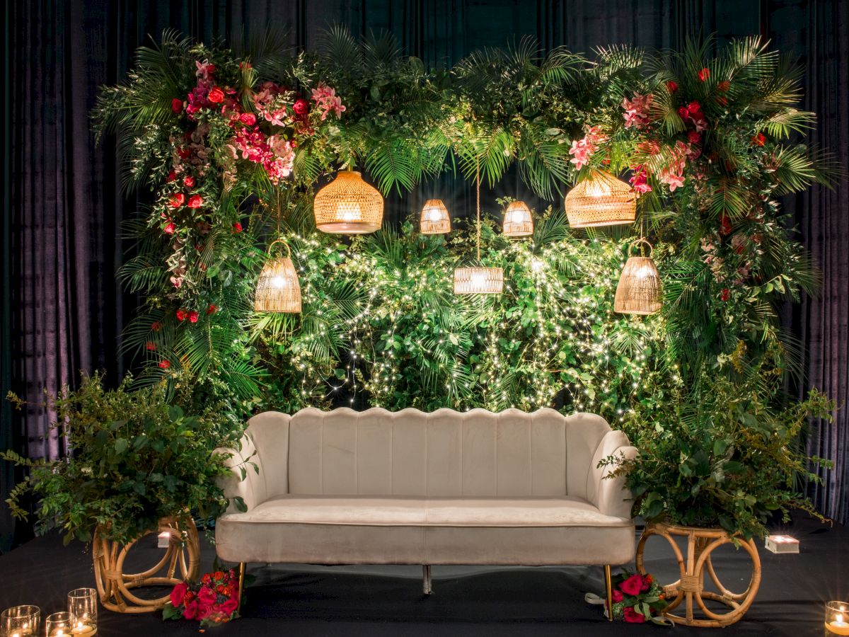 A decorated event stage with dense green foliage, pink-red flowers, warm hanging lantern lights, and a pale sofa centered.