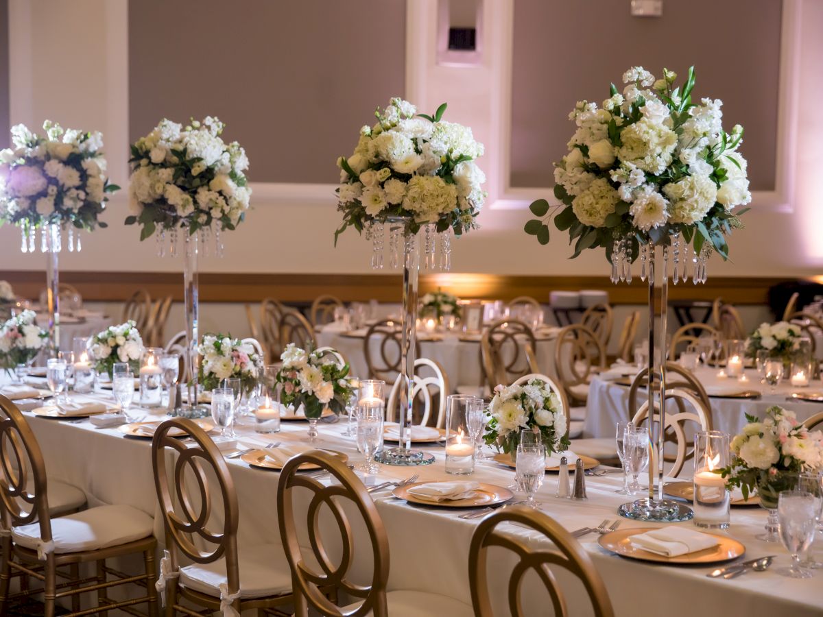 An elegant banquet setup with candlelit tables, gold chairs, and tall white-green flower centerpieces for a wedding reception.