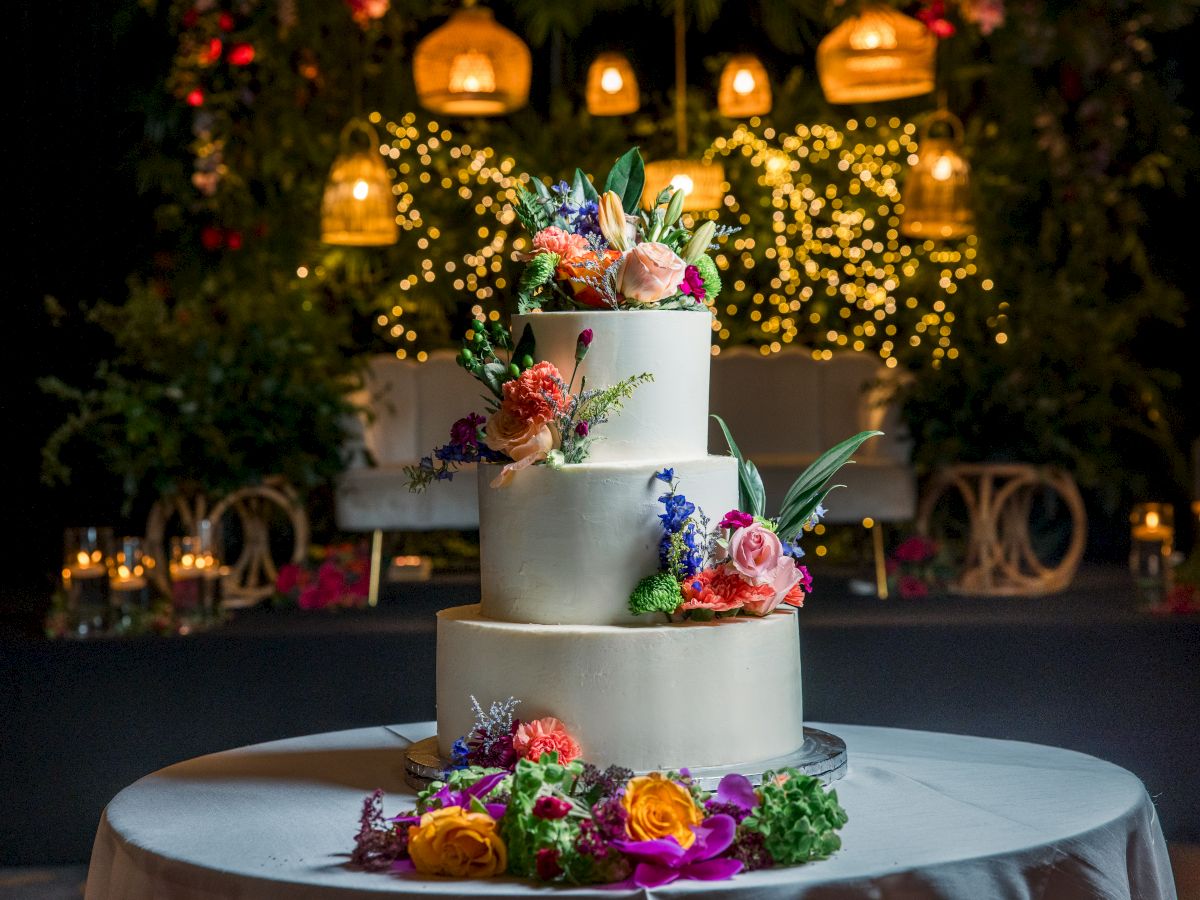 A tiered white wedding cake topped with colorful flowers, set on a table in a warmly lit garden with fairy lights.