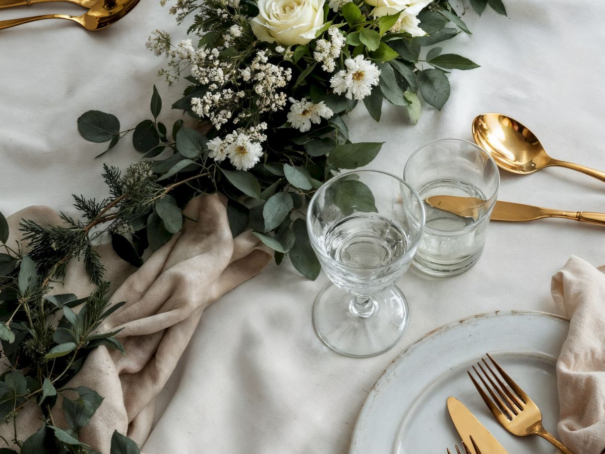 Elegant table set with white roses and greenery, gold cutlery, water glasses, and cloth napkins on a light cloth.