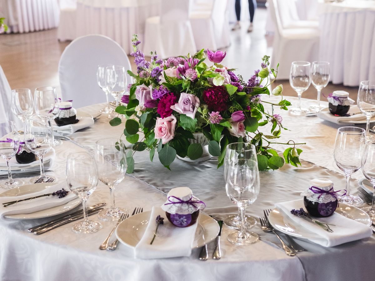 A decorated wedding-style dining table with a large purple-pink flower centerpiece, candles, and many wine glasses and cutlery on a white cloth.