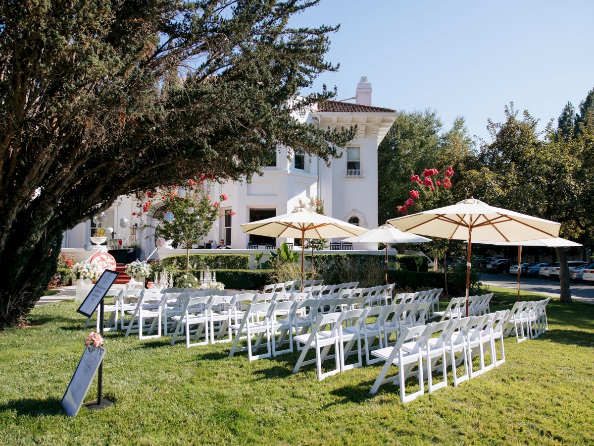 Outdoor event setup with white chairs, floral decorations, sun umbrellas, and a white house backdrop among trees.