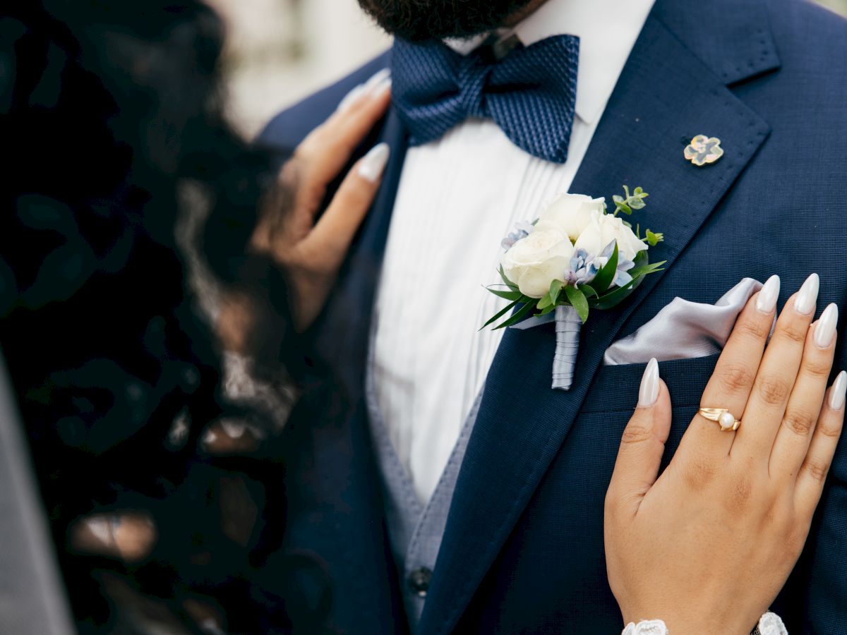A groom in a blue suit with bow tie and boutonniere is being held by a bride&rsquo;s hand with rings, in a wedding moment.