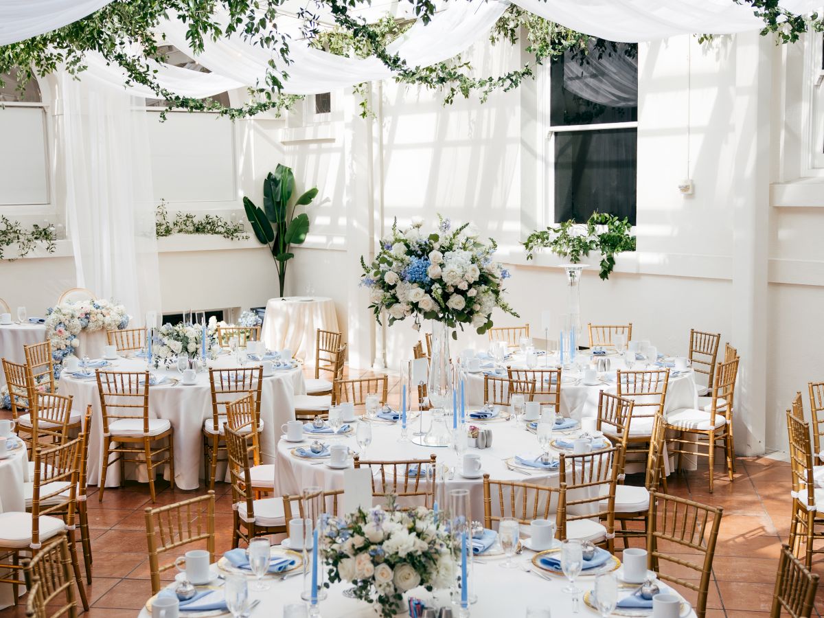 A decorated wedding reception tent with hanging greenery garlands, white tablecloths, blue accents, and gold chairs set for guests.
