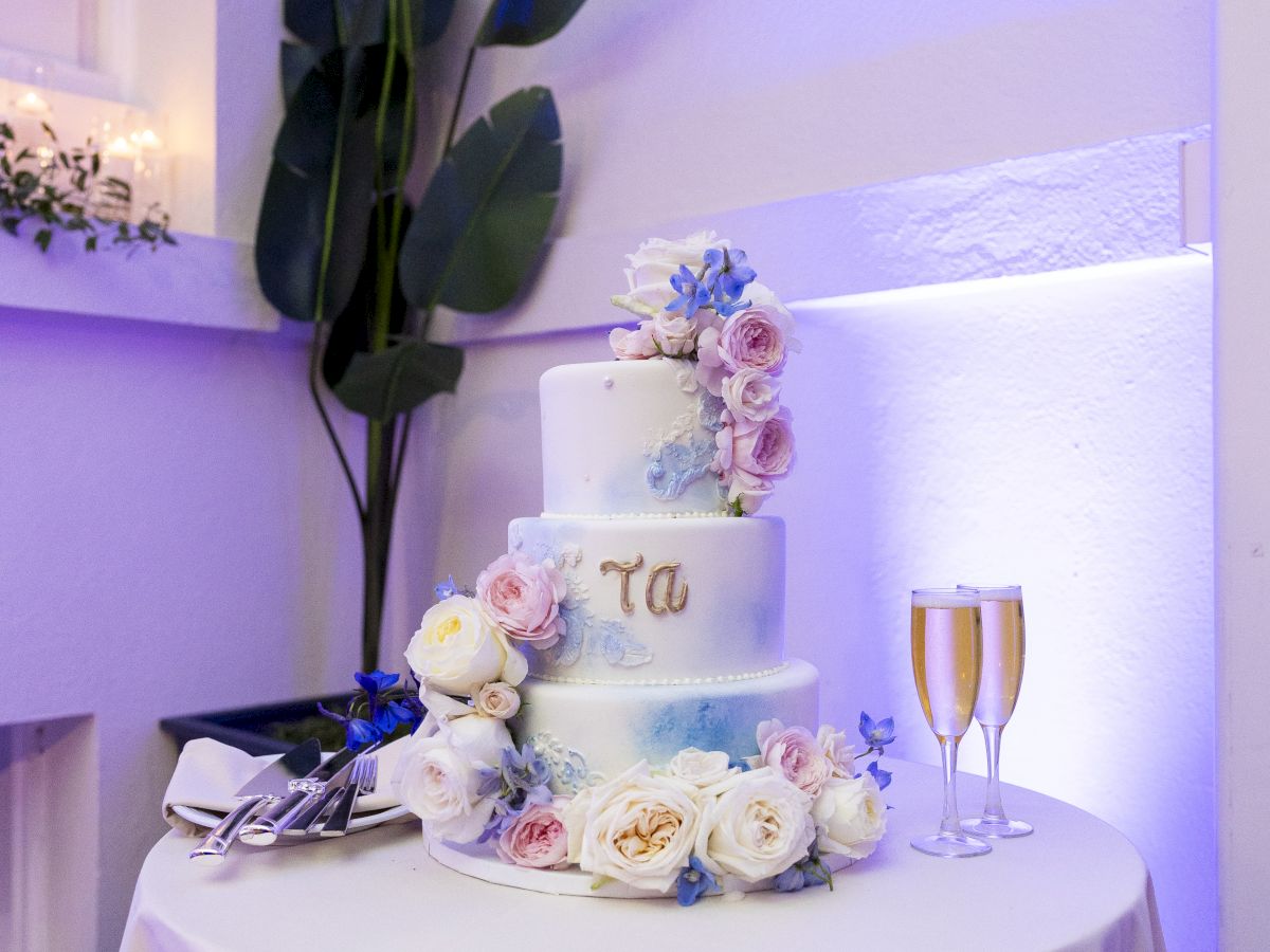 A decorated wedding cake with pastel roses and blue accents on a table, flanked by champagne flutes in soft purple lighting.