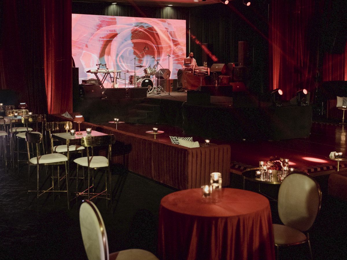 A lit stage with a drum set, music equipment, red curtains, and a large screen is seen from inside a dim event hall.