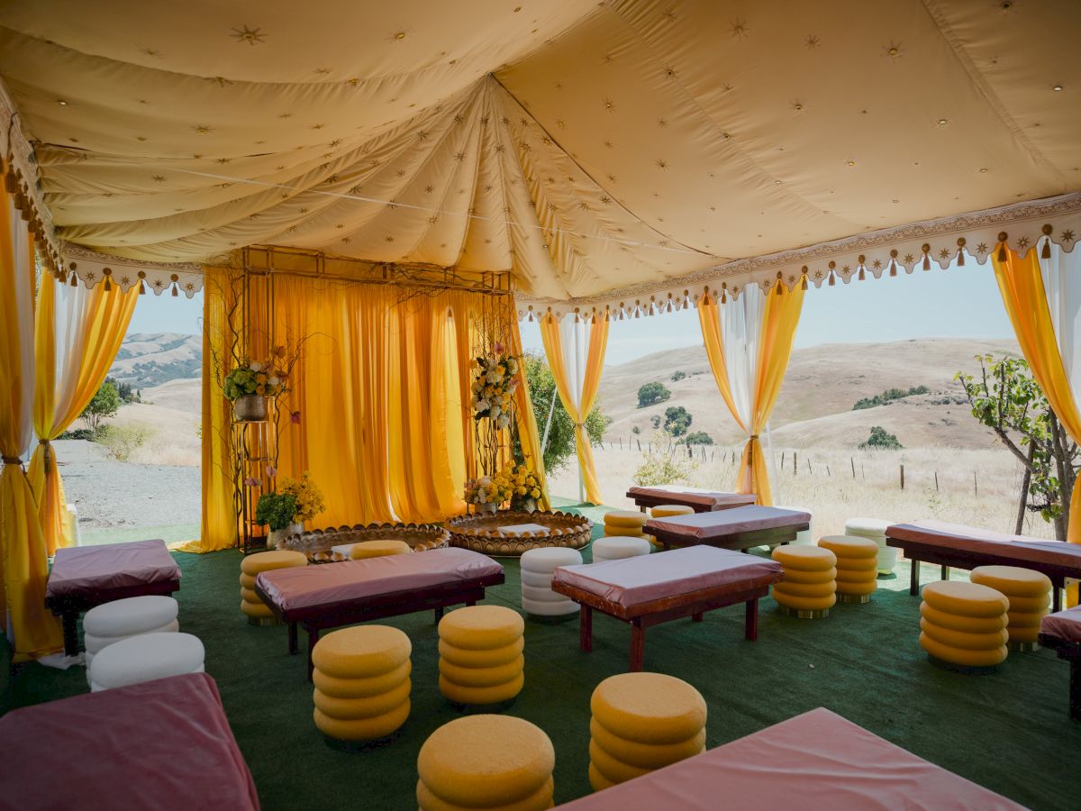 An outdoor event tent with yellow and white drapes, starry ceiling d&eacute;cor, flower arrangements, cushioned seating, and mountain views beyond.