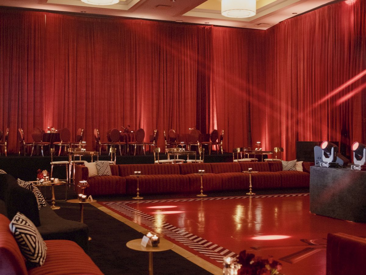 An elegant lounge with red curtains, cushioned booths, round tables, and warm ceiling lights for an event or show.