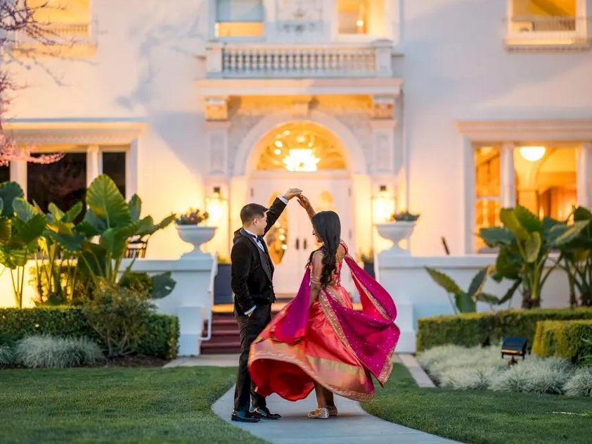 A couple in wedding attire dances and poses on a garden pathway before an illuminated building entrance at golden hour.