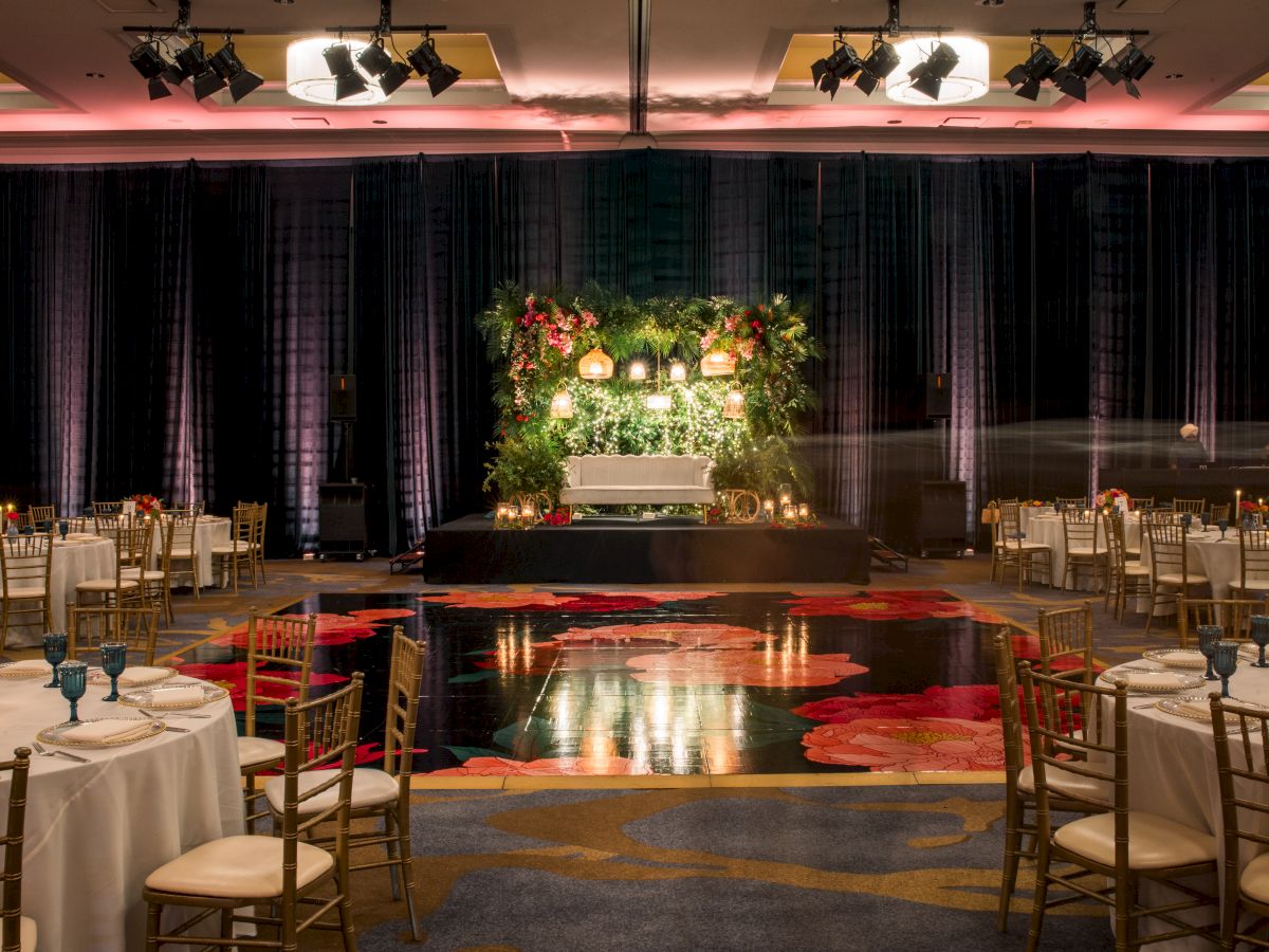 Banquet hall set for an event: floral-lit stage, black drapes, stage lights overhead, and tables with gold chairs.