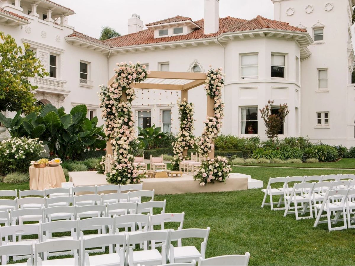 A garden wedding setup with white chairs, a floral arch backdrop, pink roses, and a decorated stage in front of a white villa.