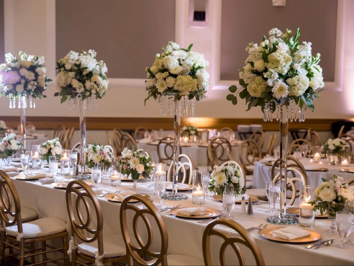 Elegant wedding reception table set with white flower centerpieces on tall stands, candles, and gold chairs in a softly lit hall.