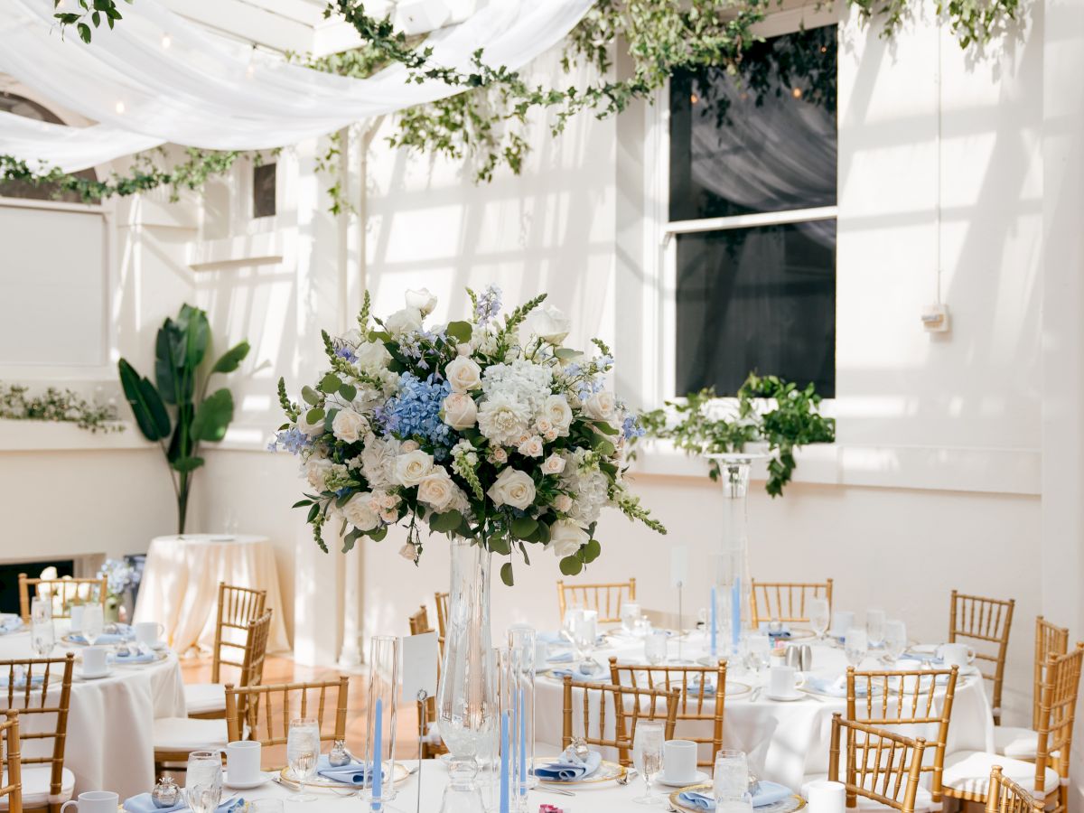 An elegant white tent reception table set with gold chairs and a tall bouquet centerpiece of white, blue, and greenery flowers.