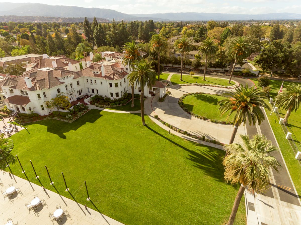 Aerial view of a luxury hillside resort with white buildings, bright green lawns, palm-lined paths, and distant mountains under sunlit skies.
