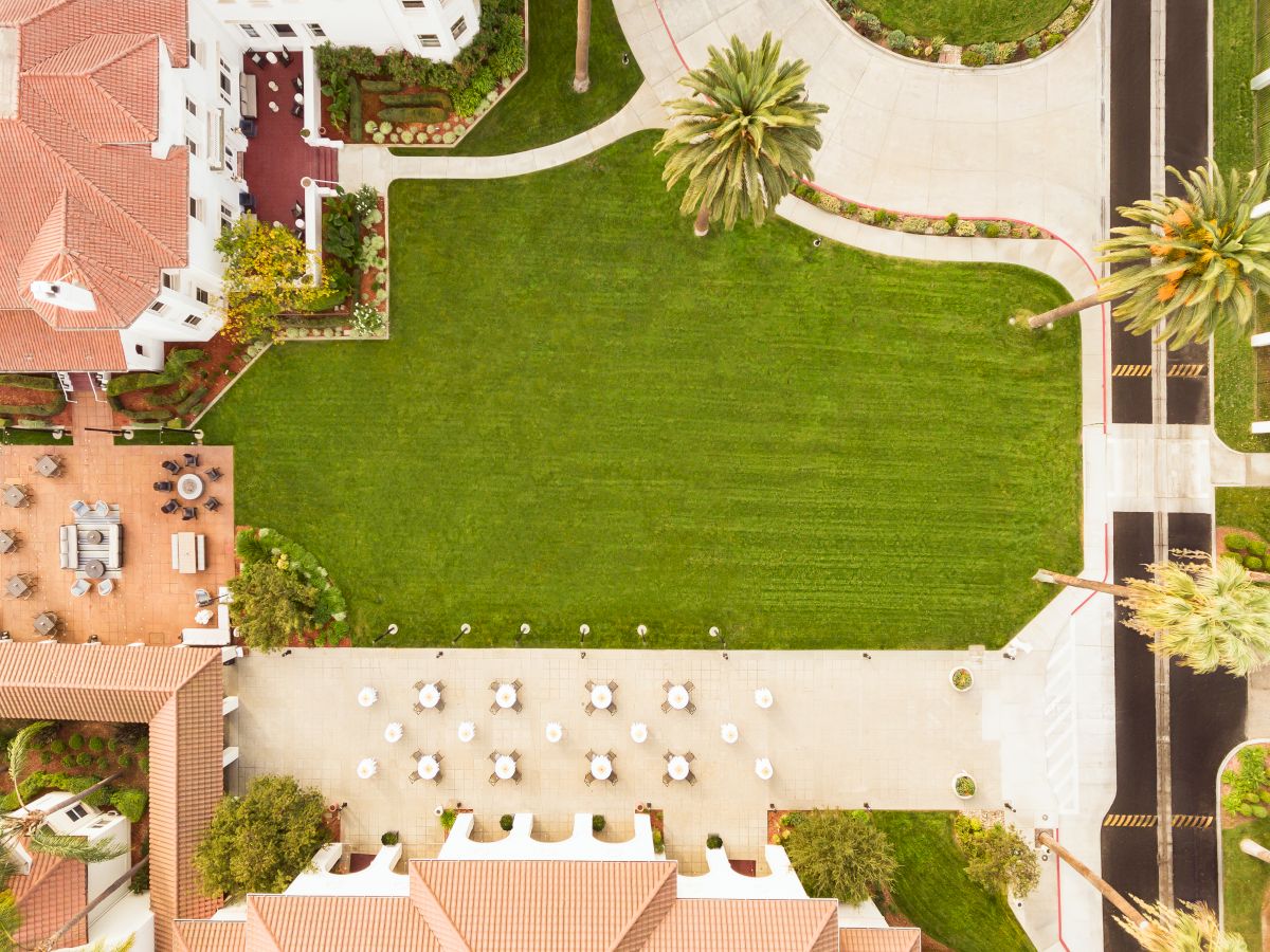 Aerial view of a resort courtyard with a large green lawn, curved paths, palm trees, and a small outdoor dining area.