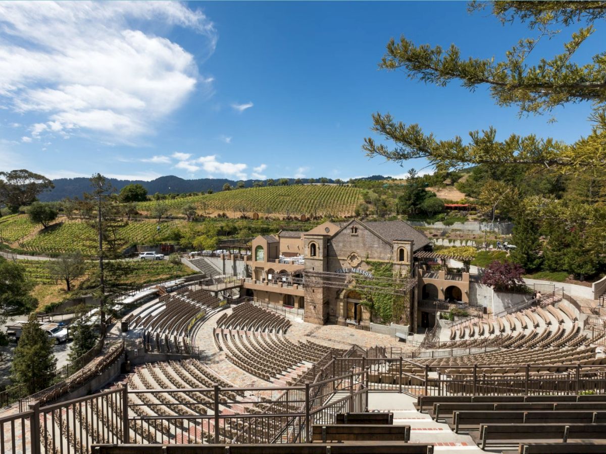 Sunlit outdoor amphitheater with rows of seats, surrounded by vineyards and trees under a blue sky.