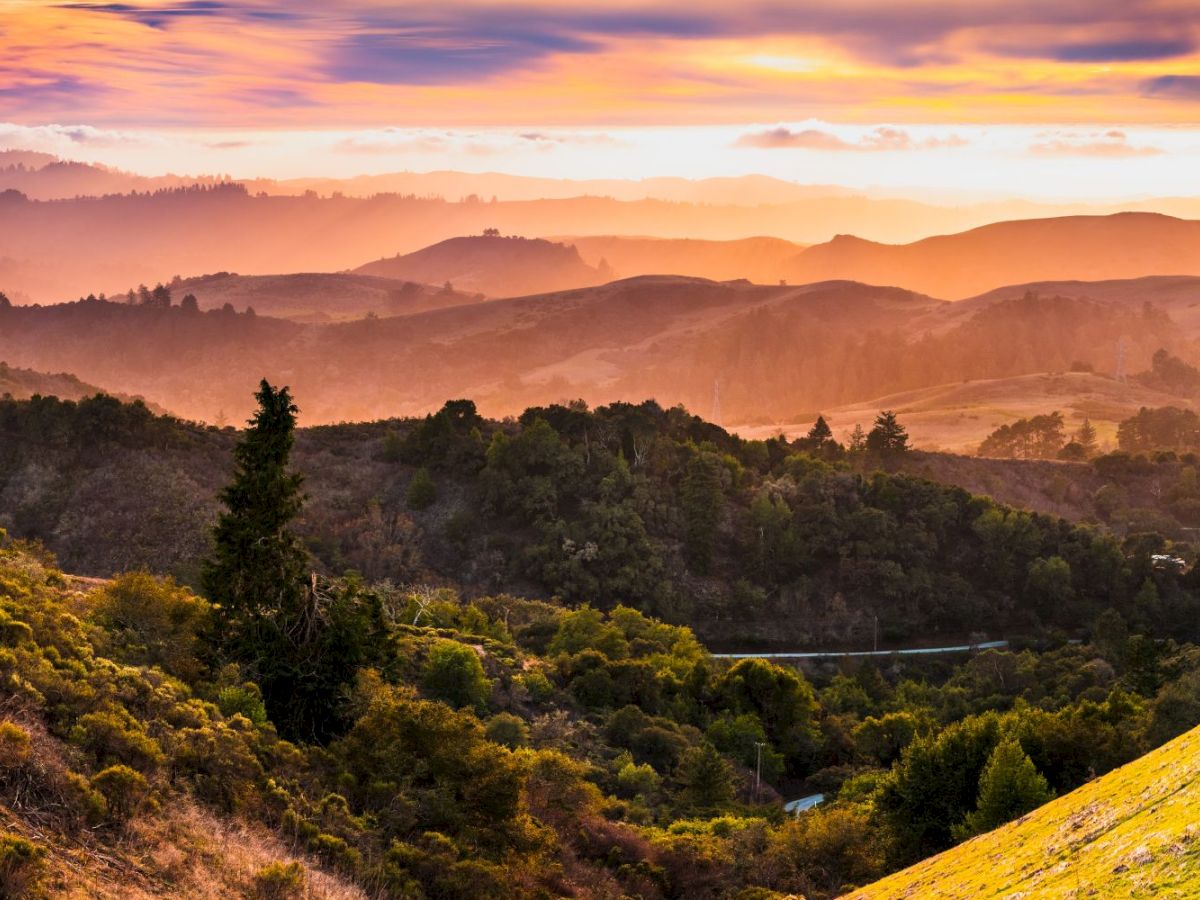 A panoramic valley at sunrise with rolling green hills, misty mountains, and warm orange/pink clouds over a winding river.