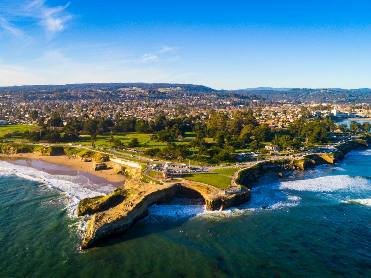 Aerial view of a sunny coastal town with a sandy beach, rocky cliffs, roads, and waves crashing along the shoreline.