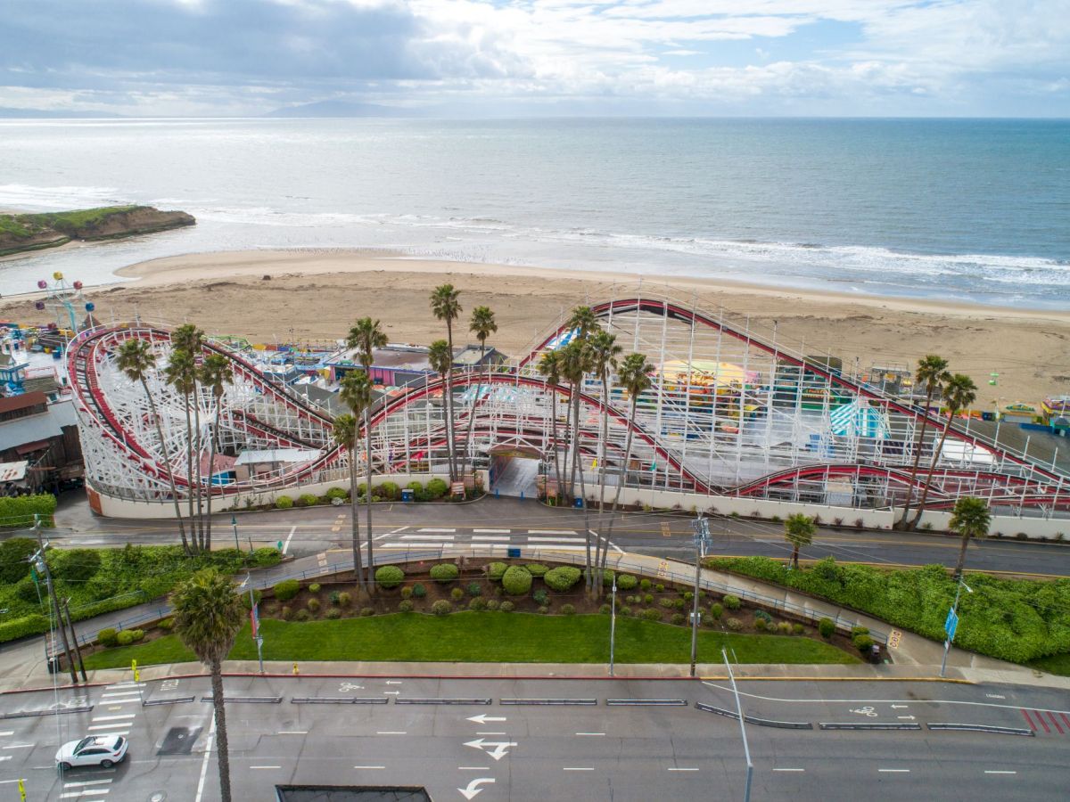 A coastal amusement park with a looping roller coaster beside a wide sandy beach and ocean under cloudy skies.