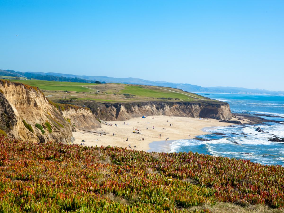 A sunny coastline with steep cliffs, a sandy beach full of visitors, ocean waves crashing below, and green hills stretching to the horizon.