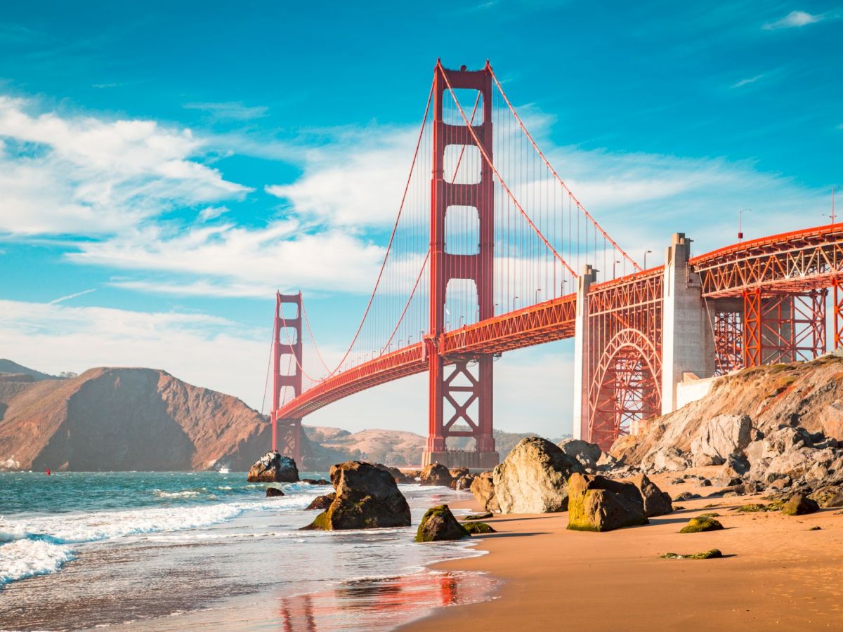 Golden Gate Bridge rises in red over the coast, with waves washing the sandy shore under a bright blue sky.