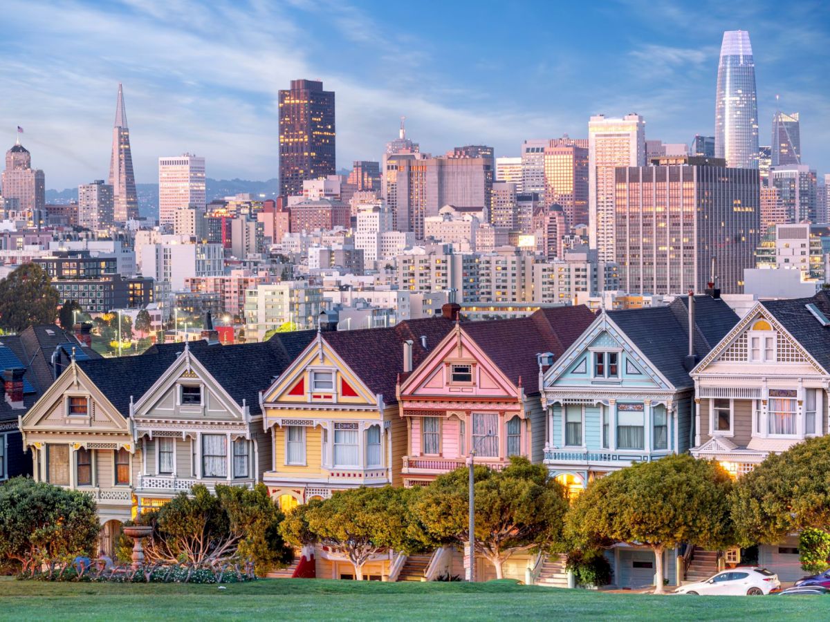 Downtown skyline at dusk with tall office towers behind colorful Victorian-style houses in the foreground.
