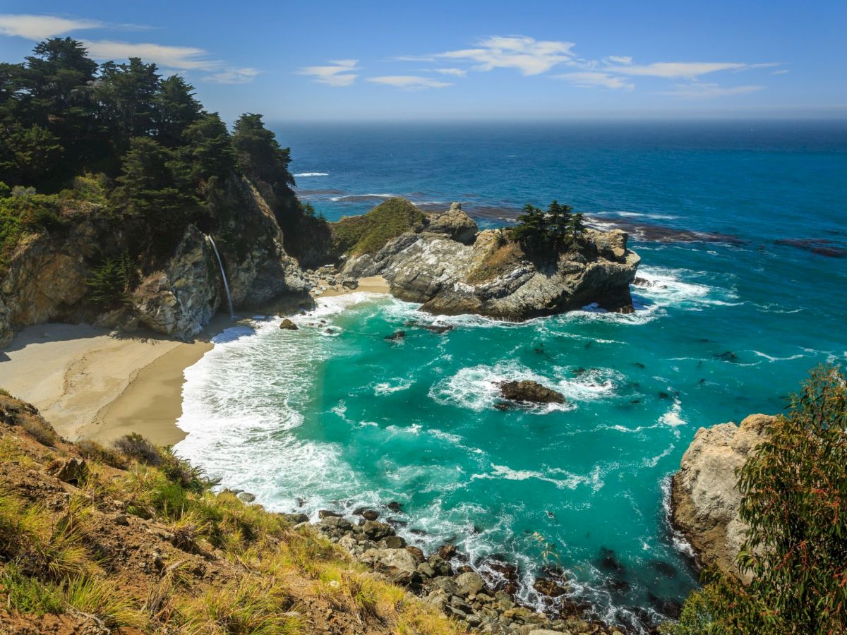 A scenic coastline with rocky cliffs, a small waterfall to a sandy cove, and turquoise ocean waves under a blue sky.