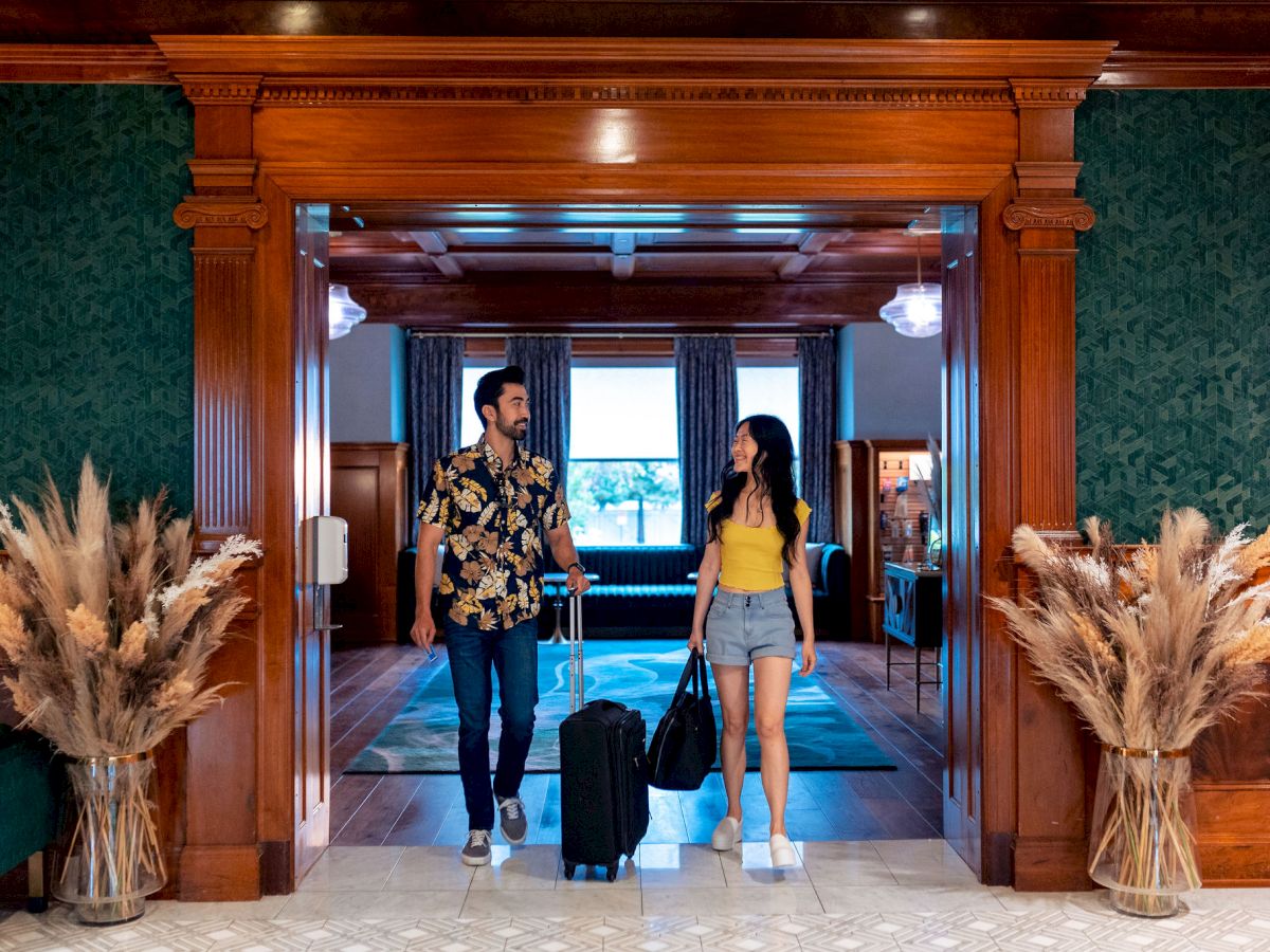 A man and woman walk into a hotel lobby holding luggage, smiling by ornate doors and fluffy pampas grass decorations.