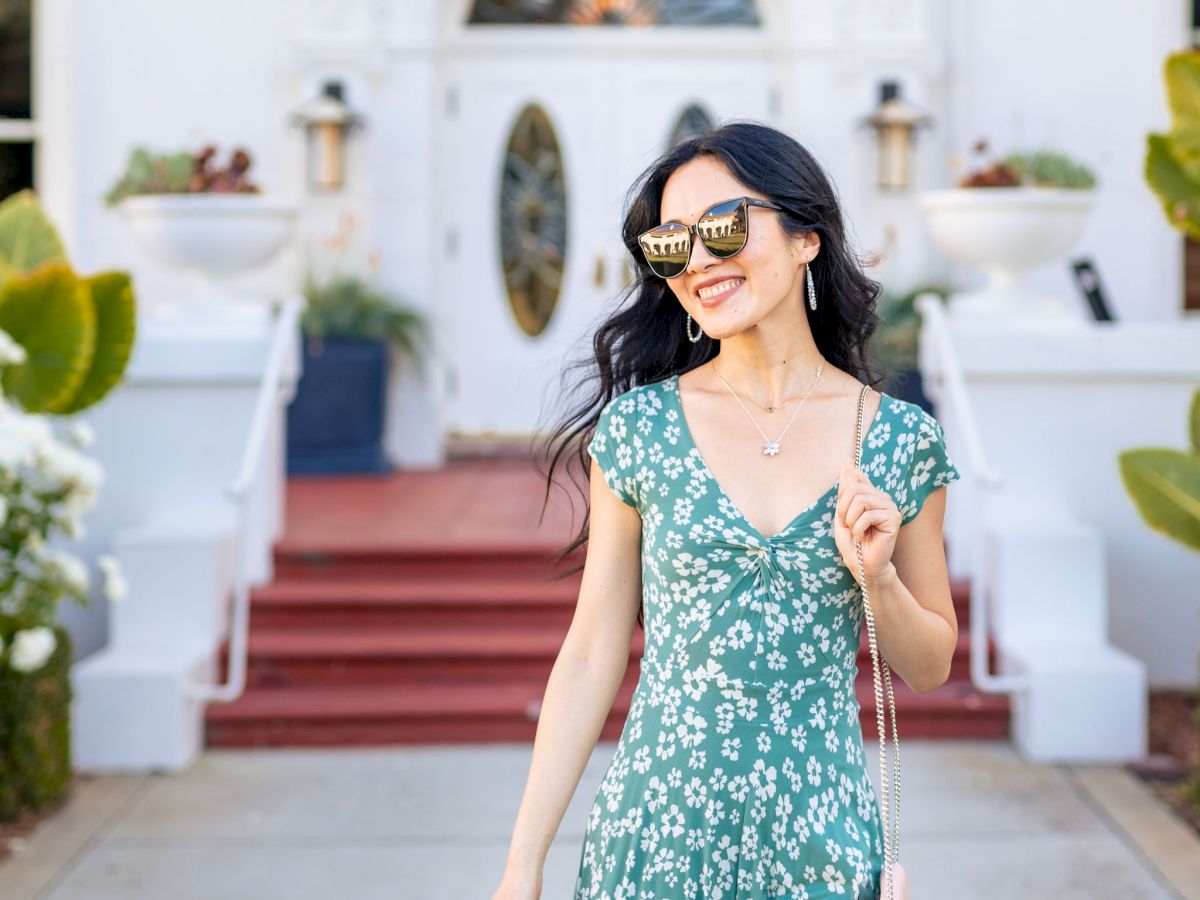 A woman in a green floral dress and sunglasses smiles outdoors in front of a white building with a big arched doorway.