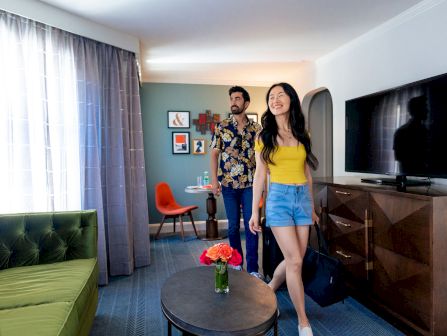 A smiling couple strolls through a modern living room, with a TV, green sofa, and a table holding colorful roses.