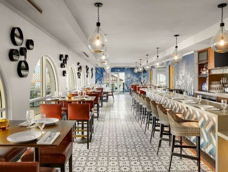 A bright restaurant dining room with long tables, patterned tile floor, pendant lights, and a bar along the wall.