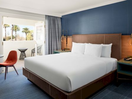 Modern hotel room with a large bed, bedside desk lamp, chair, balcony doors, and gray curtains.