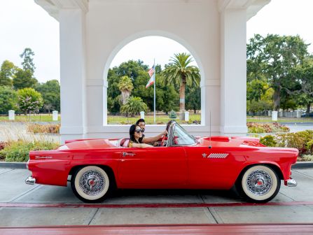 A couple rides in a red classic convertible under an archway, with an American flag and lush trees in the background.