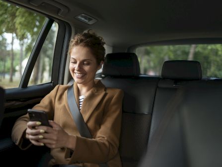 A woman in a car passenger seat smiles while looking at her phone, with trees visible through the windows.