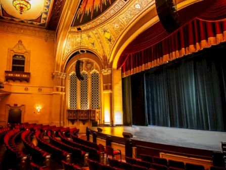 An ornate theater auditorium with red velvet seats, a grand stage, and an illuminated, painted ceiling.
