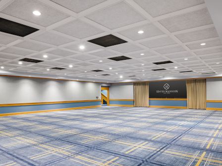 Indoor event hall with acoustic ceiling, blue carpet, and a stage with gold curtains plus a &ldquo;Hayes Mansion San Jose&rdquo; sign.