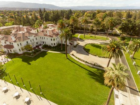 Aerial view of a sunny resort estate with white villas, manicured lawns, palm trees, winding paths, and distant mountains.
