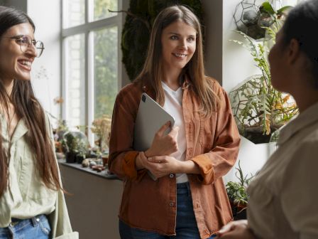 In a plant-filled indoor shop, two women chat while one smiles and holds a tablet, surrounded by hanging greenery.