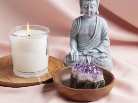 A lit candle sits beside a small Buddha statue and a bowl holding an amethyst crystal on a wooden tray.