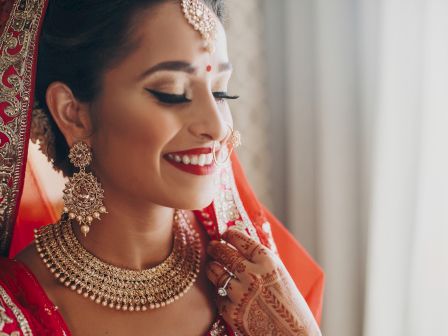 A woman in traditional red attire, smiling in side profile, wearing ornate jewelry, sindoor, and henna, with a bright bindi.