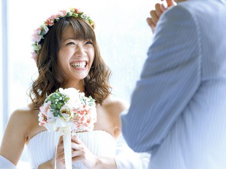 A smiling bride in a floral crown holds a pastel bouquet while a groom in a blue suit stands beside her, celebrating love.