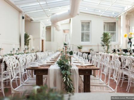 A bright wedding reception hall with white chairs, a decorated central table, greenery, and candleholders.
