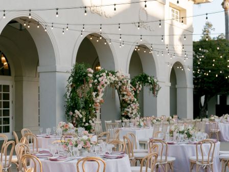 Outdoor wedding setup with white arches, hanging string lights, and banquet tables dressed with floral centerpieces and candles.